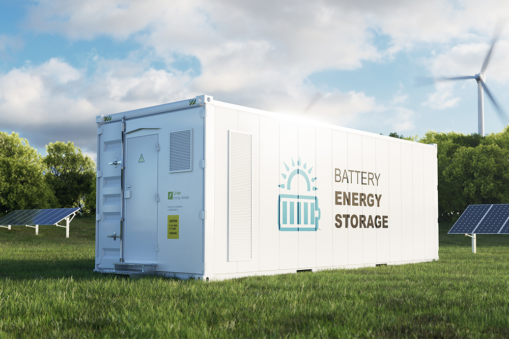 A  white battery energy storage unit sits in a green field, with solar panels and a wind turbine in the background.
