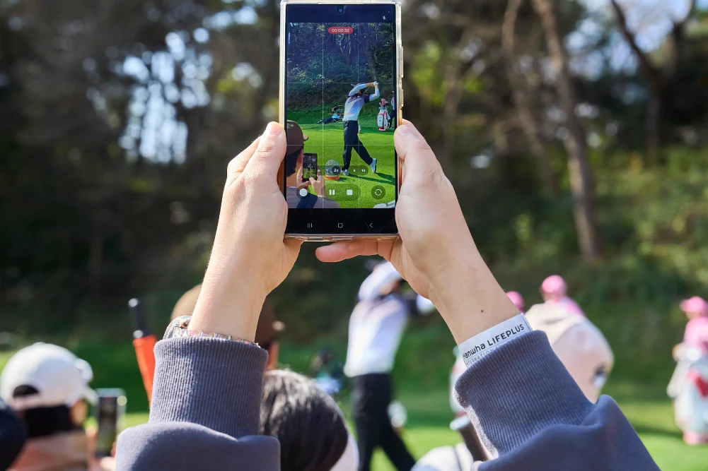 A spectator films a golfer’s swing on a smartphone.