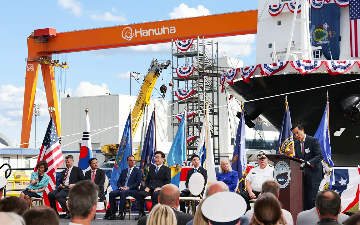 Several leaders sit in front of Hanwha Philly Shipyard in front of an audience during a christening ceremony.